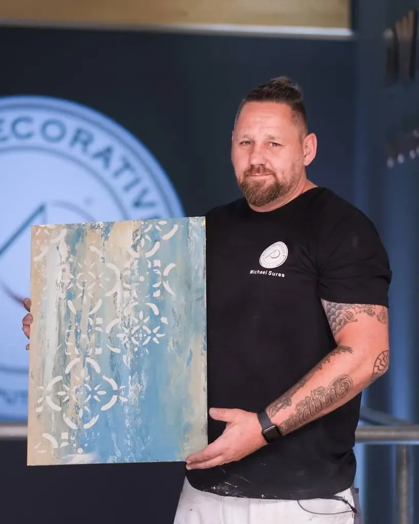 Michael Sures, Inhaber von Eigenwerk, hält eine Musterplatte mit türkis-heller Schablonenstruktur in die Kamera