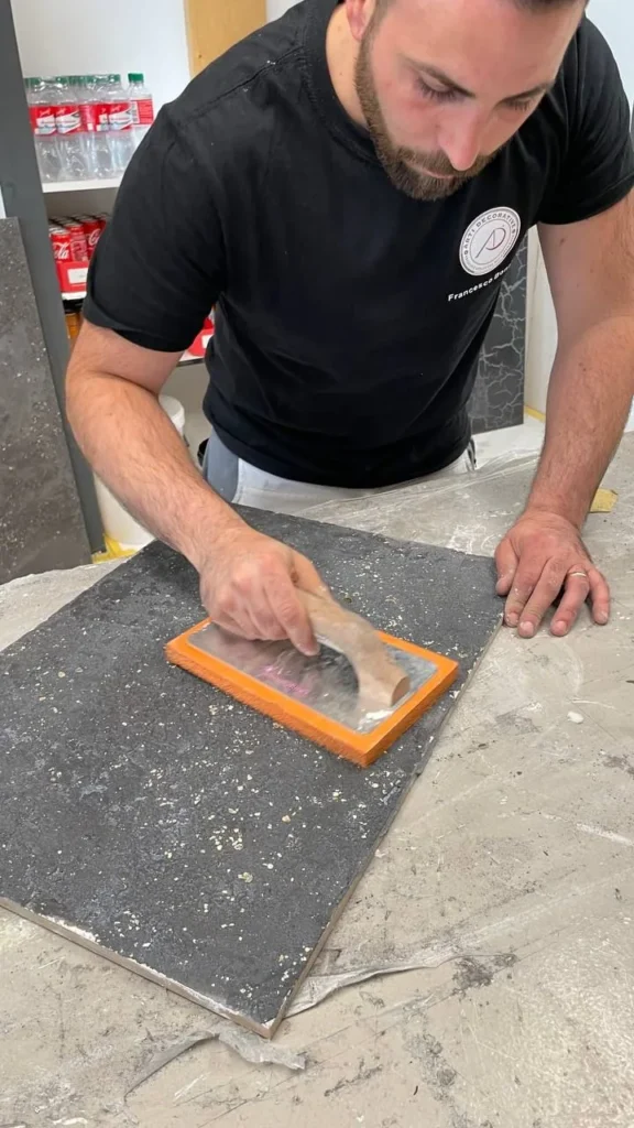 Mitarbeiter zieht mit der Glättkelle eine dunkle Spachtelmasse über eine Musterplatte