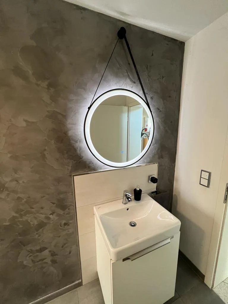 Gäste-WC mit fugenlos gespachtelter Wand und rundem Spiegel mit Hinterleuchtung